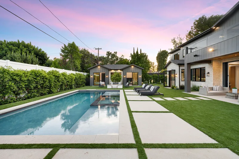 Long rectangular lap pool beside a modern house at sunset, with concrete pavers set in artificial turf and loungers by a pool house