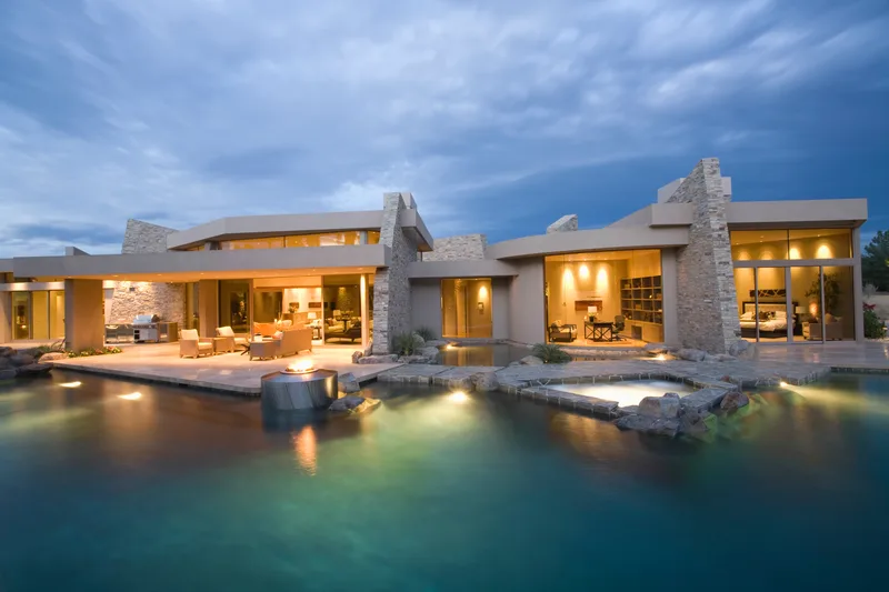 Modern stone house lit up at dusk behind a dark freeform pool with a lit spa