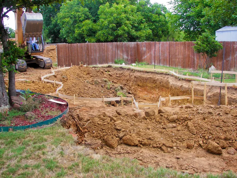 Freshly dug pool shape in a fenced backyard, with wood forms set and an excavator parked behind