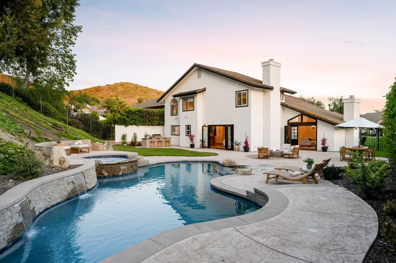 Freeform pool with a raised stone spa spilling into it at dusk, beside a white house on a hillside