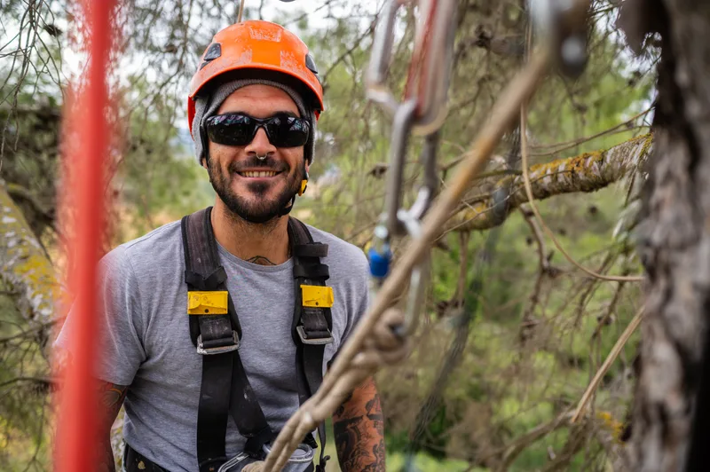 Caleb Whitlock, smiling arborist in an orange climbing helmet, sunglasses and harness, beside his climbing ropes