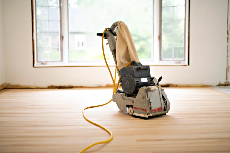 Drum floor sander standing on freshly sanded pale hardwood in an empty, sunlit room