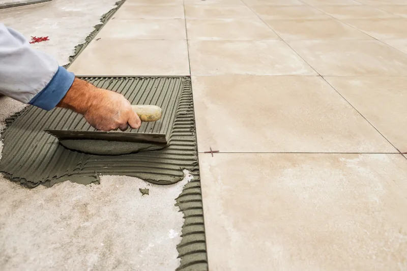 Tile setter spreading thin-set with a notched trowel beside rows of large beige floor tiles