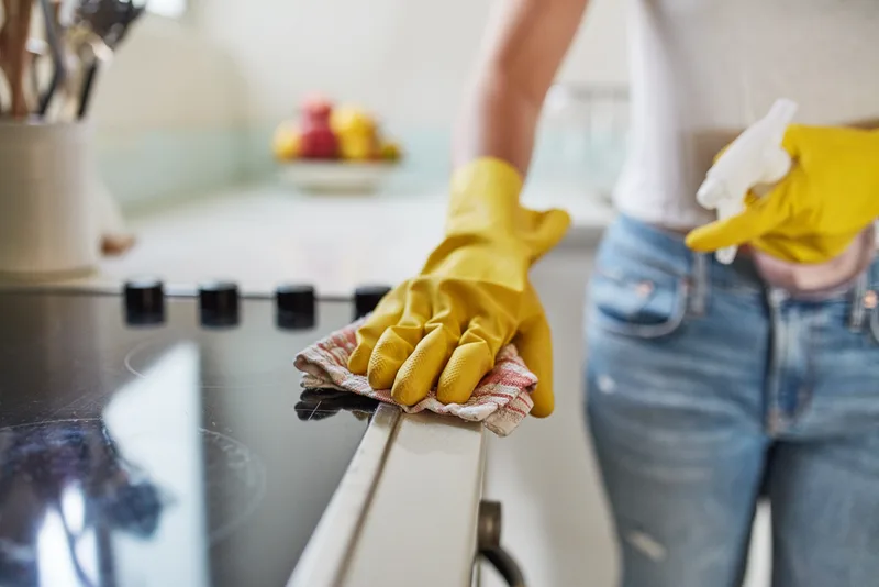 Close-up of yellow-gloved hands wiping down a kitchen range edge with a cloth, spray bottle in the other hand