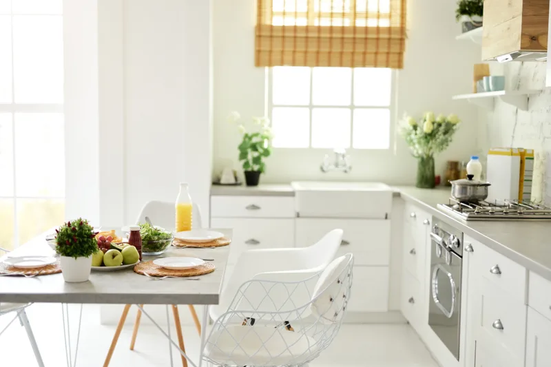 Bright white kitchen with a farmhouse sink, fresh flowers on the sills, and a breakfast table set with fruit