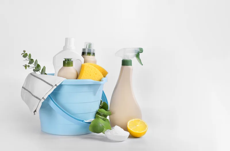 Blue bucket packed with gentle cleaning supplies — pump bottles, a sponge, fresh lemon and limes, and a sprig of eucalyptus