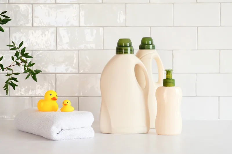 Refillable bottles of gentle cleaning product beside a folded white towel and two rubber ducks on a white-tiled bathroom shelf