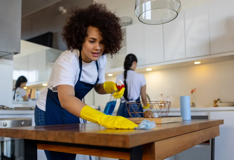 Three Tidy House cleaners working through a kitchen together, the crew lead wiping a wooden dining table in yellow gloves