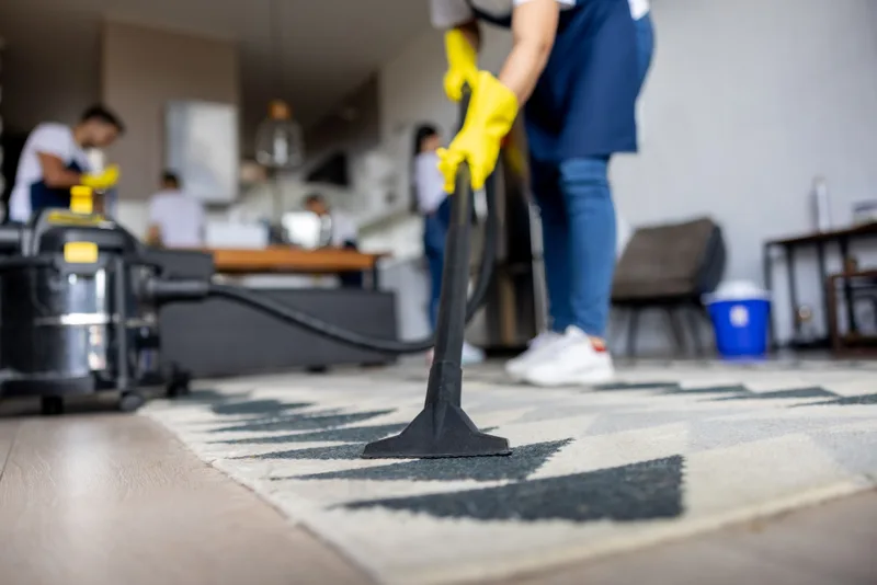 Cleaner running a vacuum wand over a black-and-white patterned area rug while teammates work in the kitchen behind