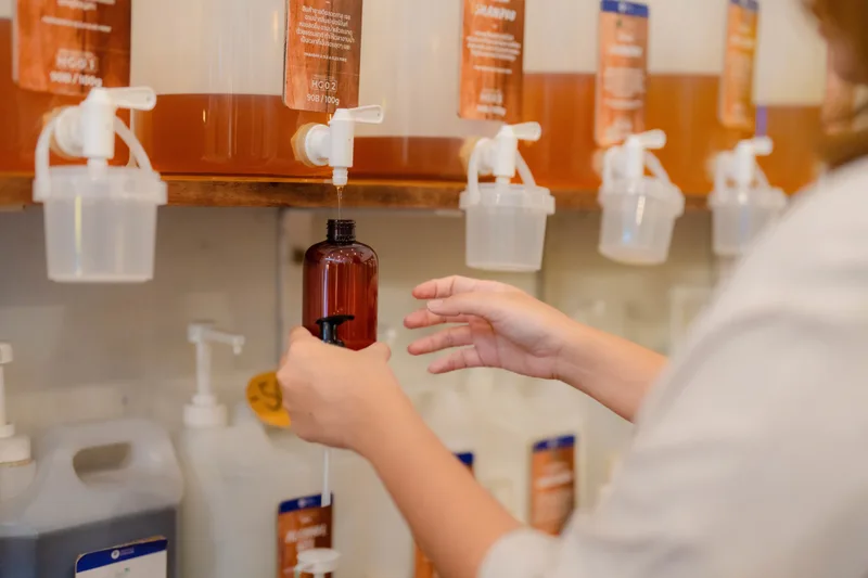Hands refilling an amber bottle from a bulk dispenser at a zero-waste cleaning-supply refill station
