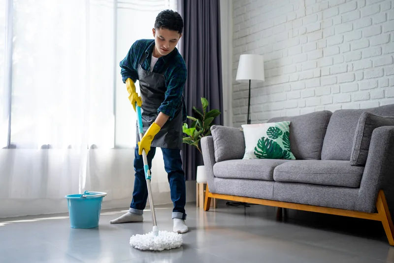Tidy House cleaner in an apron and yellow gloves mopping the floor of a bright living room beside a grey sofa