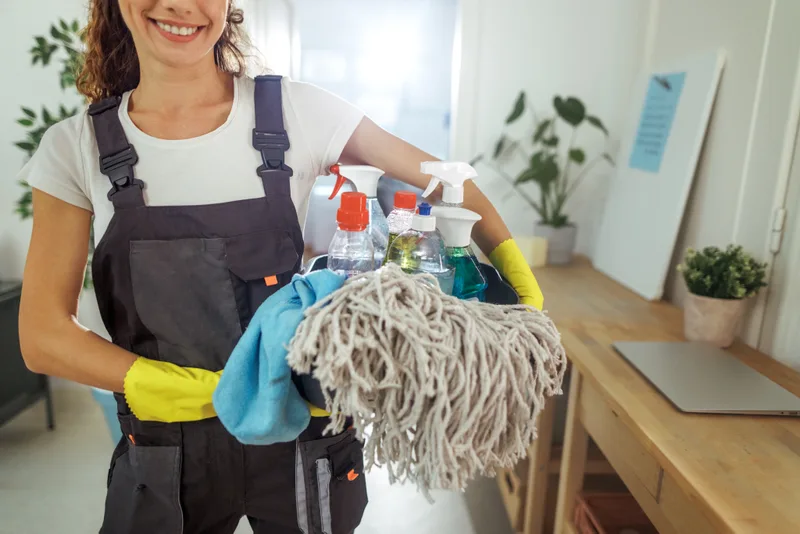 Smiling Tidy House crew member carrying a caddy of refillable cleaning supplies and a fresh mop head down a sunlit hallway