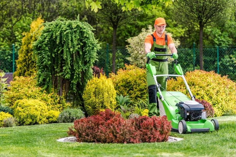 A lawn technician in a green uniform mowing a lush lawn beside trimmed shrubs and flower beds