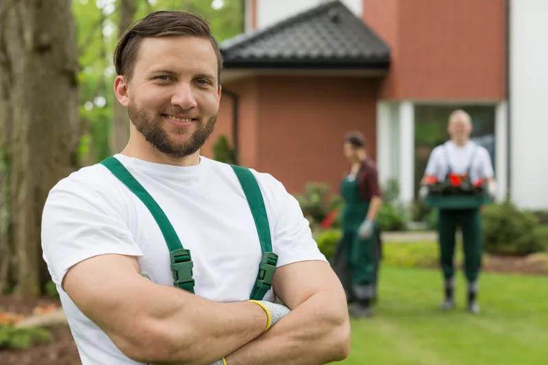 A smiling Tuft crew member in green overalls standing on a lawn, with teammates working behind him