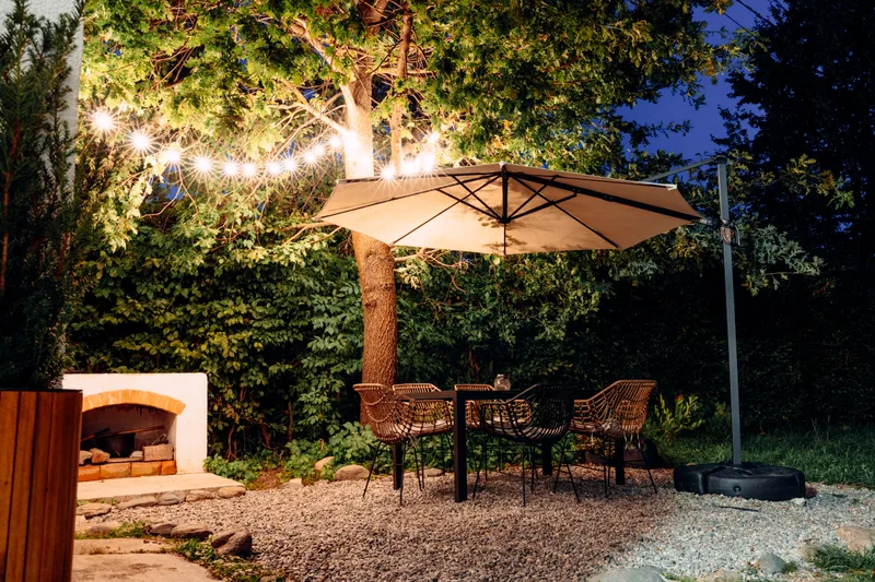 A gravel dining terrace at dusk under string lights, with rattan chairs, a cream umbrella and a white outdoor fireplace