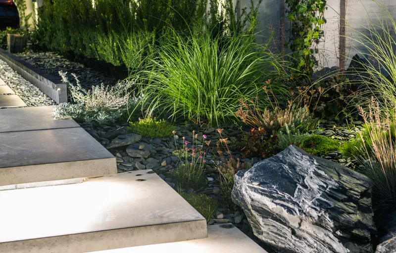 Pale concrete steps floating over dark slate chippings beside a granite boulder and tall ornamental grasses
