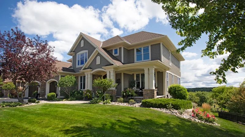 Traditional grey two-storey home with layered foundation planting and a stone-edged lawn in full leaf