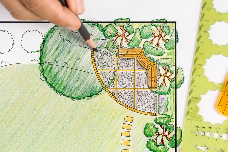 A designer's hand drawing a coloured landscape master plan with pencil and scale rulers