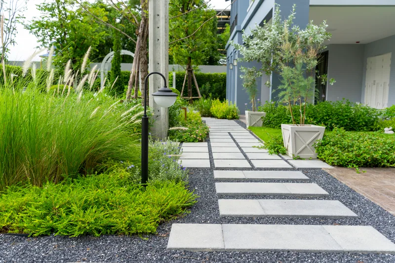Pale stepping pavers set in dark gravel, leading past ornamental grasses and a black lamp post to a modern grey house
