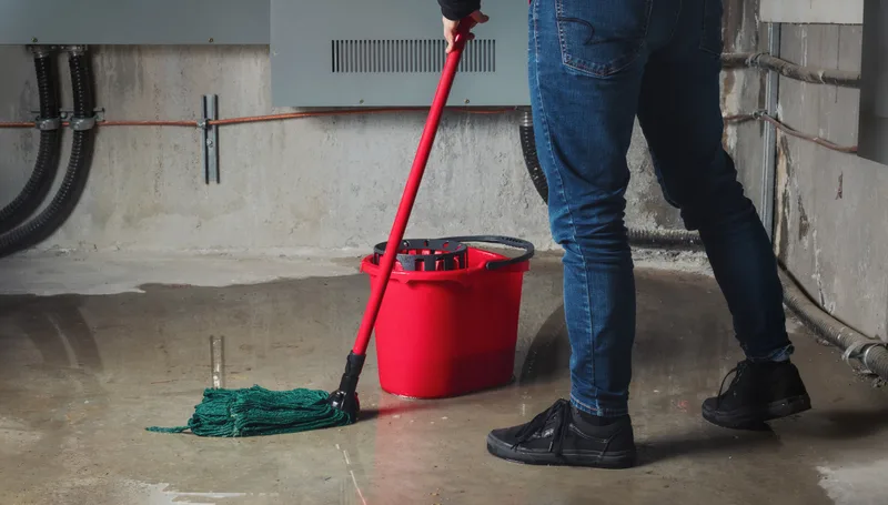Someone mopping standing water from a concrete basement floor beside a red bucket