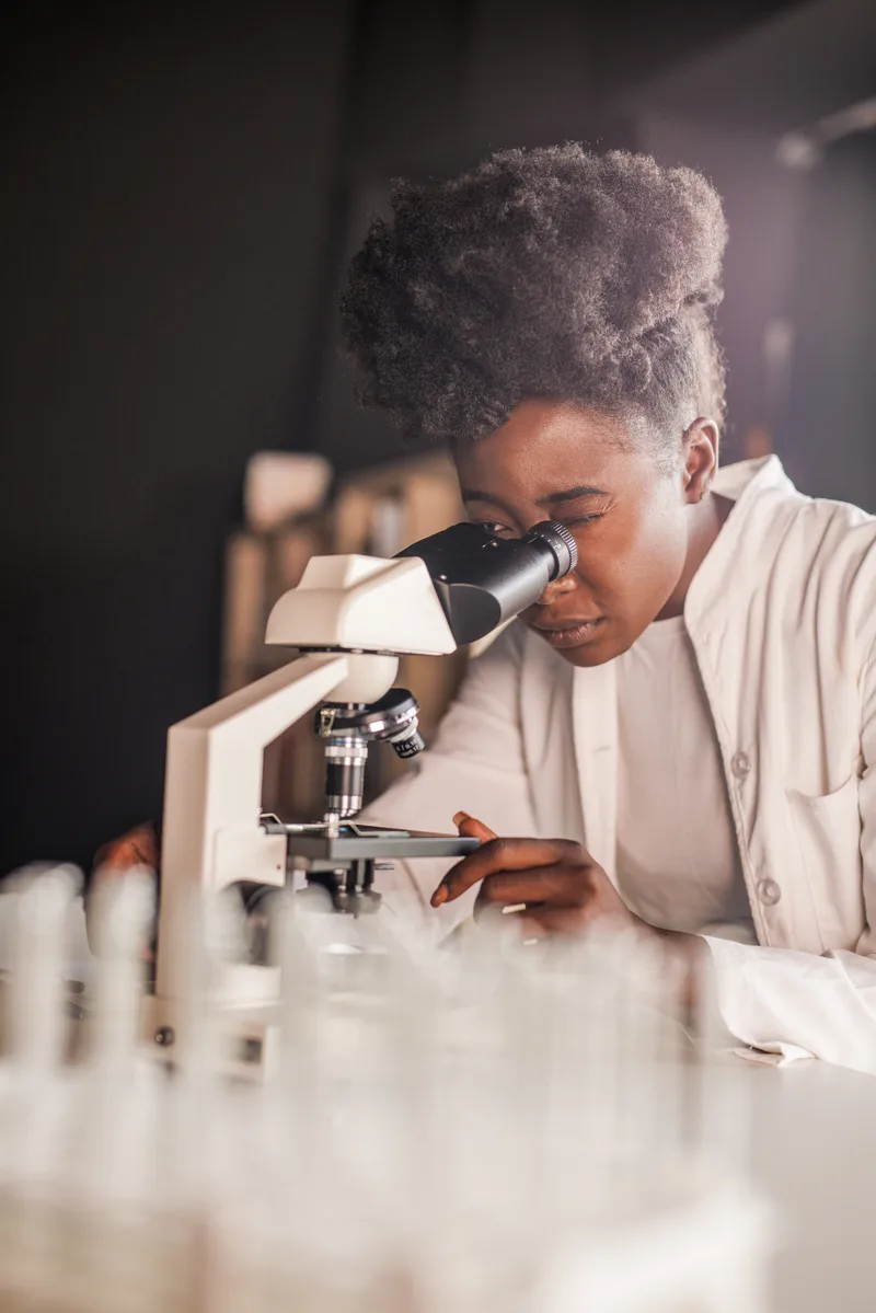 A lab analyst in a white coat looking through a microscope at a bench of sample tubes