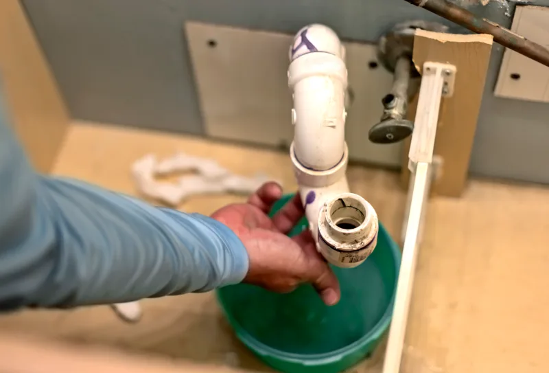 A person checking the drain trap under a bathroom sink for a slow leak, with a bucket underneath