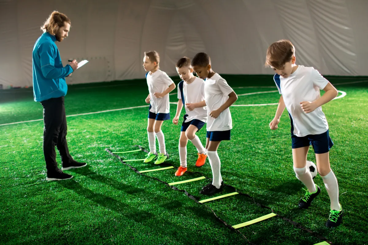 Youth sports coach guiding a small group of kids through an agility ladder drill on an indoor turf field