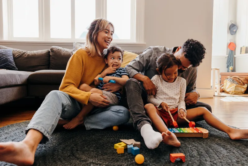 A mother and father sit on their living room floor laughing with their two young children