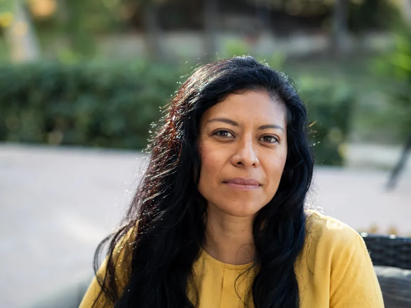 A woman in a yellow sweater sits outdoors in the sun with a calm smile
