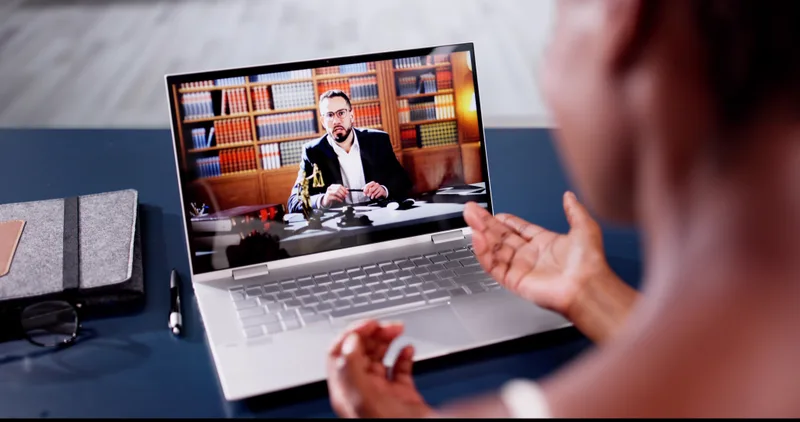 A client on a video call with a lawyer, seen over her shoulder on a laptop screen
