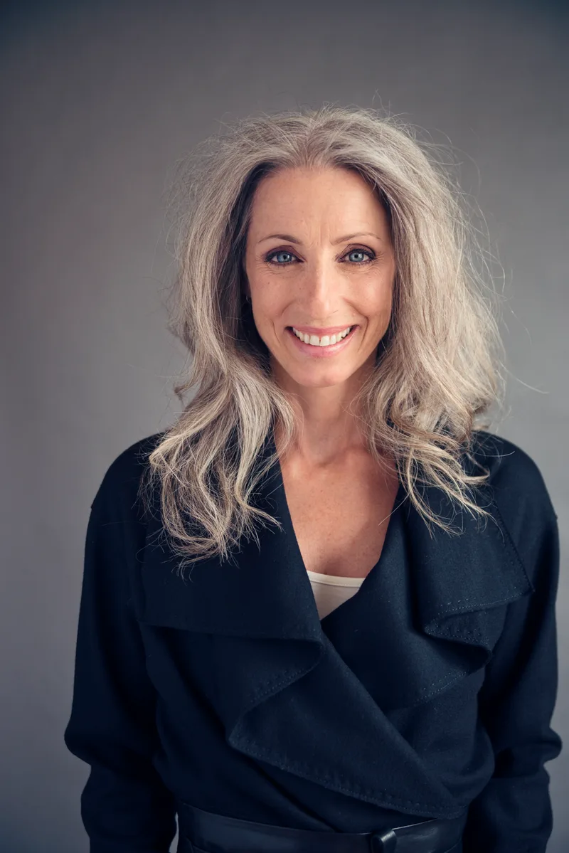 Claire Whitfield, a smiling woman with long silver hair in a dark jacket, photographed against a grey background