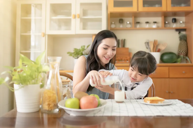 A mother pours a glass of milk for her young daughter at a bright kitchen table