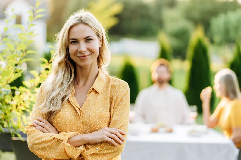 A woman smiles with her arms folded at a relaxed family dinner in a sunlit garden