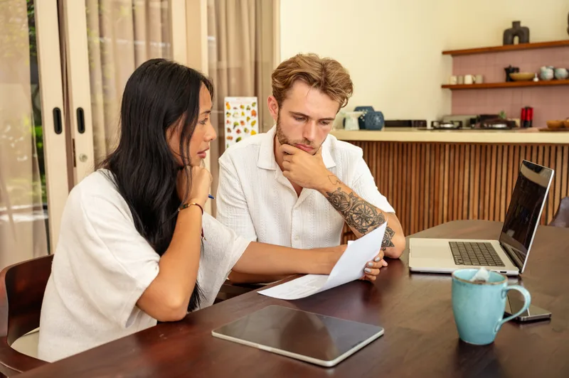 A young couple reads through paperwork together at their kitchen table, with a laptop and a mug beside them