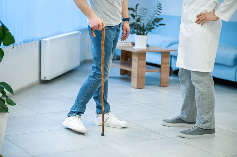A patient walking with a cane meets a doctor in a bright clinic hallway