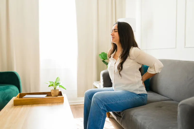 A woman sitting on her sofa presses a hand to her lower back in pain