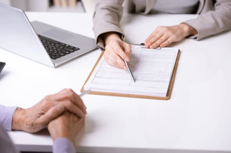 An advisor points to a line on a form while an older client fills it in at a desk