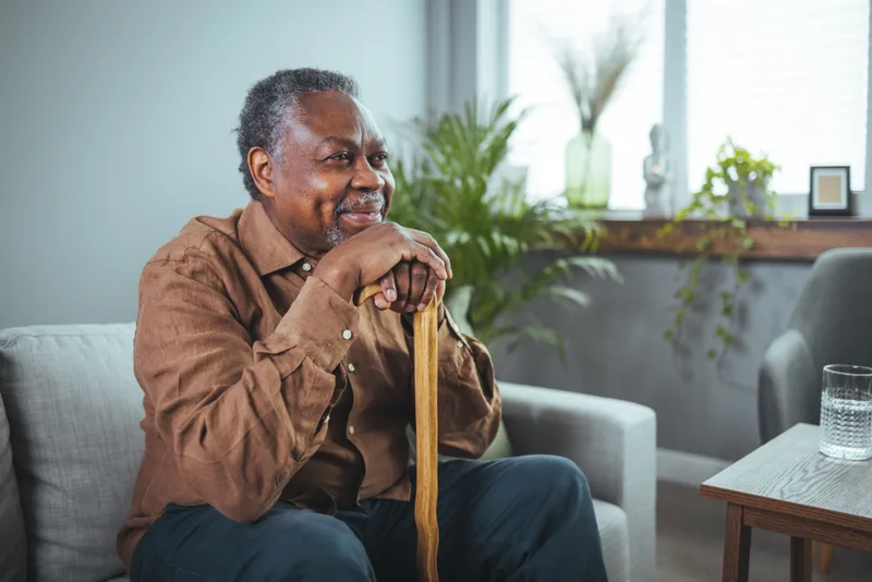 A smiling older man rests both hands on his wooden cane in his living room
