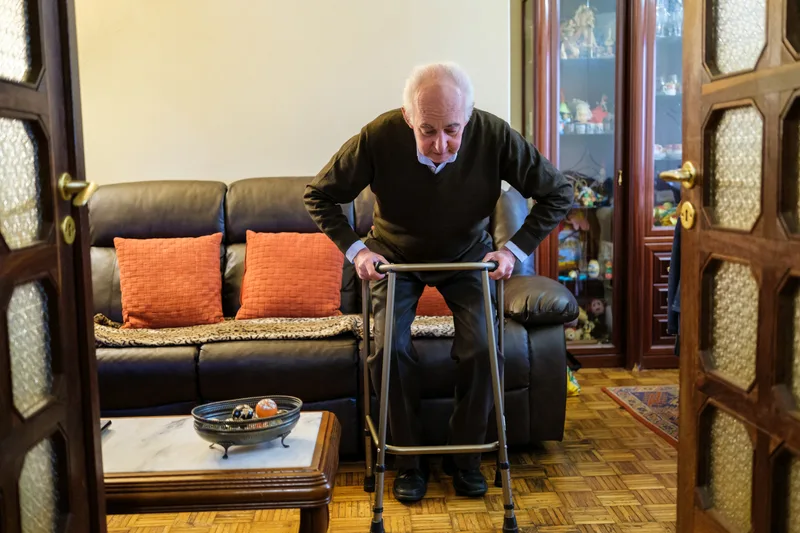 An older man uses a walker to stand up from his sofa at home