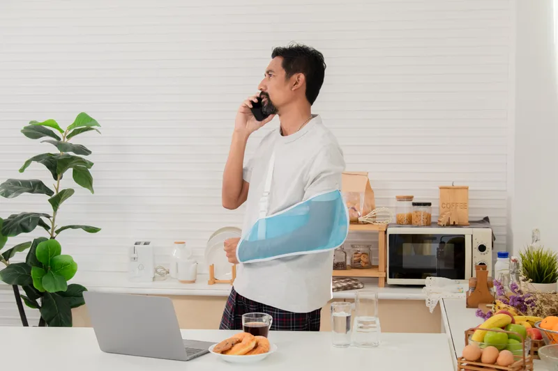 A man with his arm in a sling talks on the phone in his kitchen while off work
