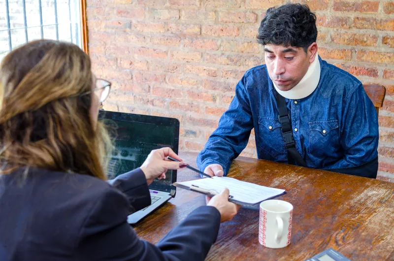 A lawyer walks an injured worker wearing a neck brace through a document at a meeting table