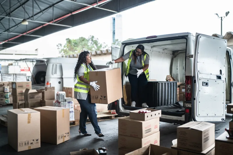 Two workers in safety vests load boxes into a delivery van at a loading area