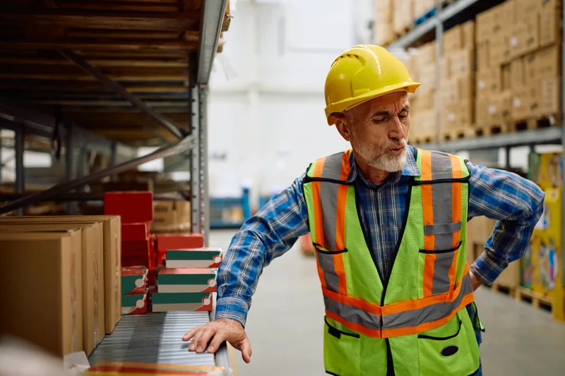 An older warehouse worker in a hard hat and safety vest steadies himself on a shelf, one hand pressed to his lower back