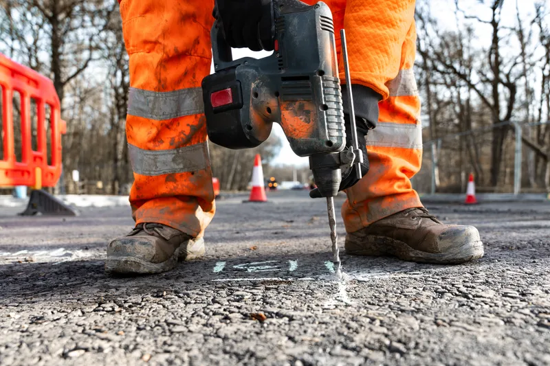 A road crew worker in orange work clothes and safety boots runs a heavy breaker on asphalt