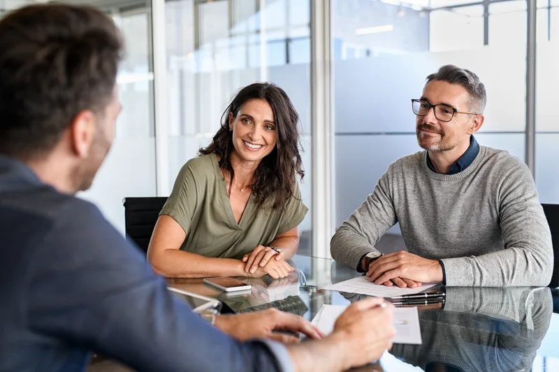 Two people sit across a glass table from a lawyer, smiling and talking through their options