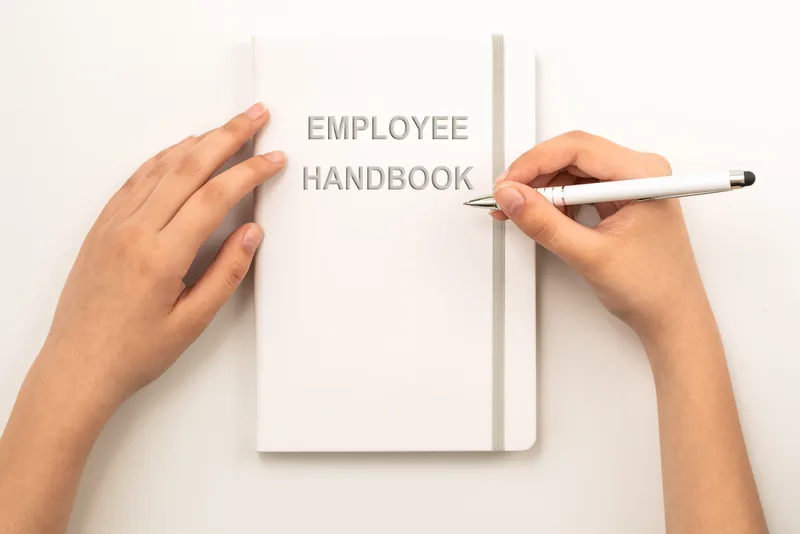 Hands open a white employee handbook and hold a pen above it