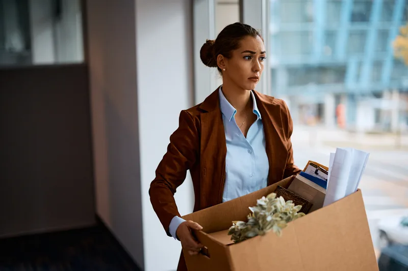 A woman in a brown blazer carries a box of her belongings out of an office building, looking ahead