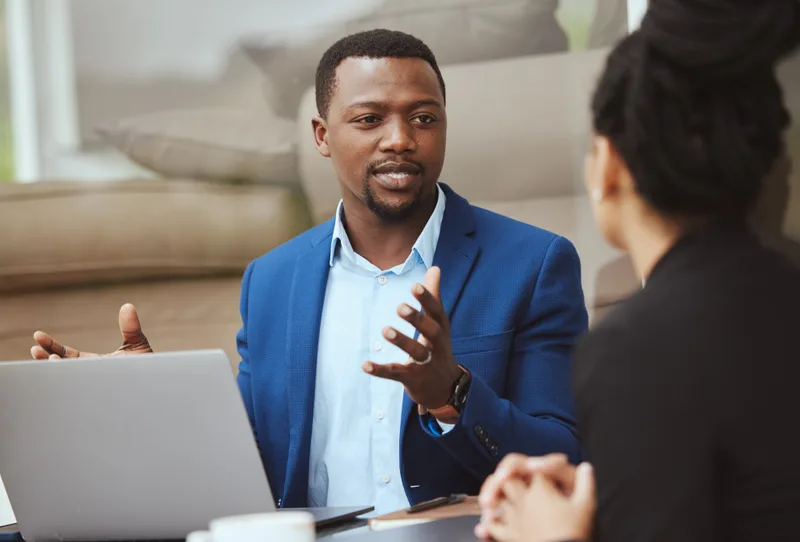 A financial adviser in a blue blazer talking through figures with a client across a laptop