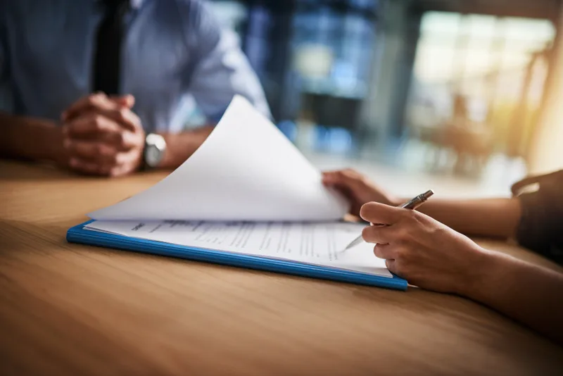 Two people at a wooden table, one turning the pages of a stapled document on a blue clipboard