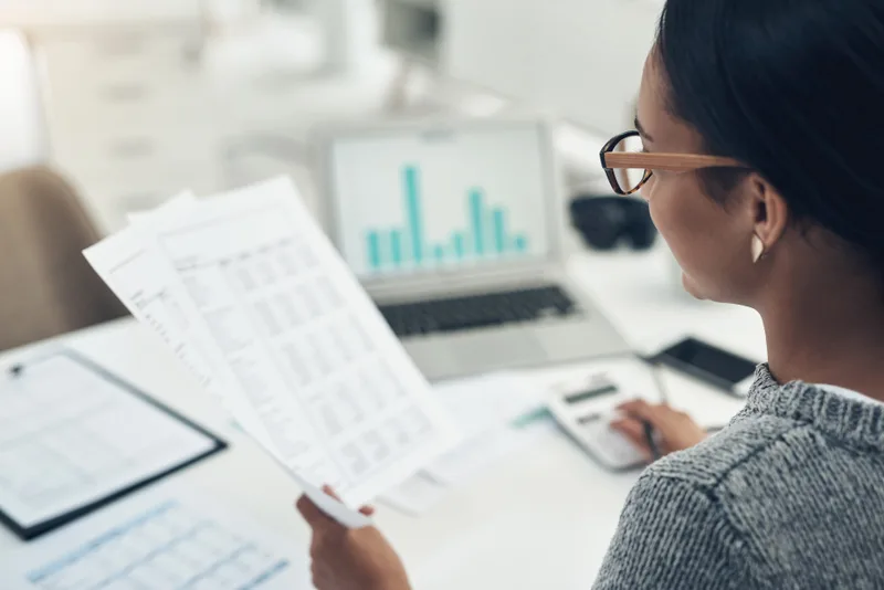 Woman at a desk reading printed financial statements beside a laptop showing a bar chart